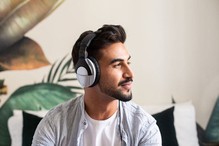 Happy ethnic guy in headphones smiling and looking away while listening to music at home