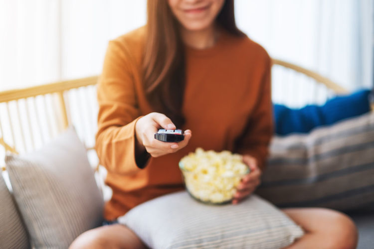 A woman eating pop corn and searching channel with remote control to watch tv while sitting on sofa