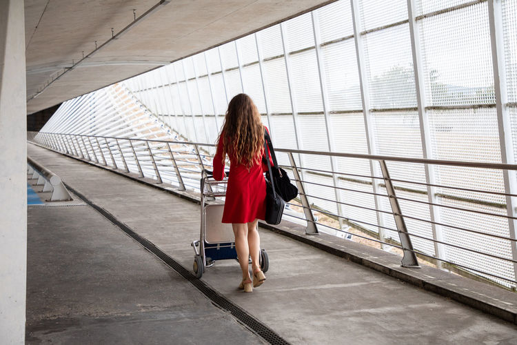Back view of traveling female walking in airport with baggage trolley and waiting for flight