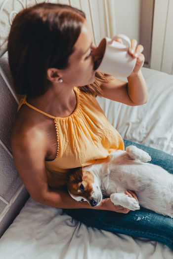 High angle view of young woman with dog drinking coffee in cup while sitting on bed
