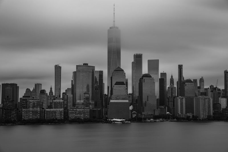 Modern buildings in city by river against cloudy sky