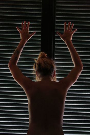 Rear view of naked woman with arms raised standing by window