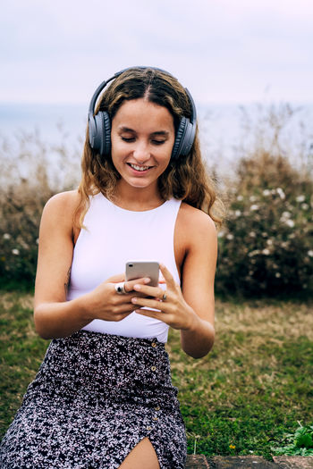 Enthusiastic woman in headphones placing on brick ledge in grass and listening to music from mobile phone