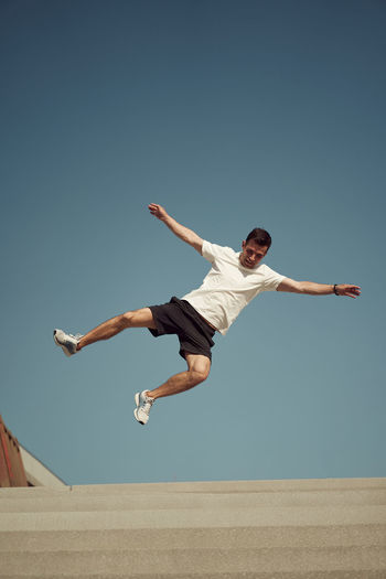 From below of active male jumping high and showing parkour stunt against blue sky in summer