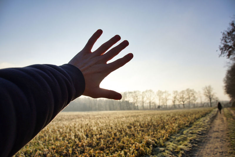 Cropped hand of man blocking sunbeam on field against sky