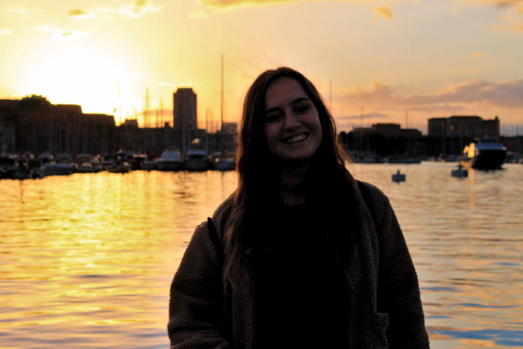 Portrait of smiling woman standing at sea against sky during sunset