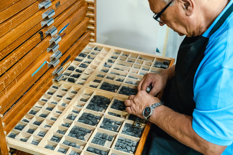 From above of crop concentrated senior male artisan in apron and eyeglasses choosing printing press letters from wooden box during work in traditional atelier