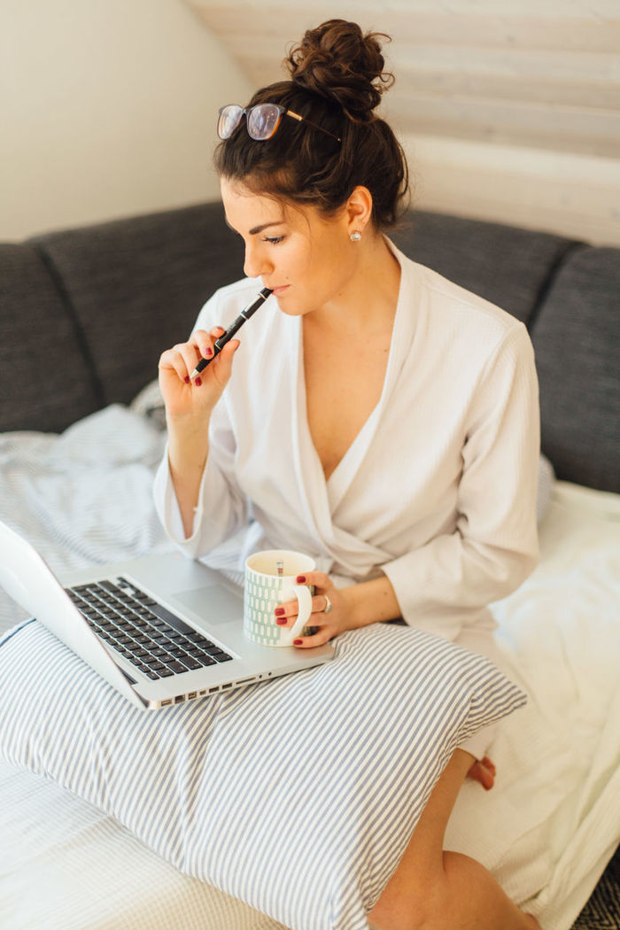 Young woman holding coffee cup using laptop while sitting on bed at home