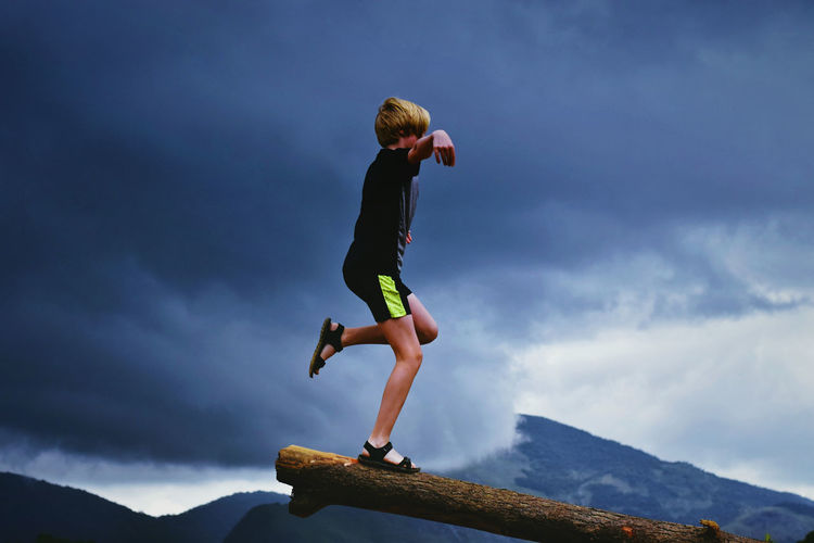 Side view of boy standing on log by mountains against cloudy sky