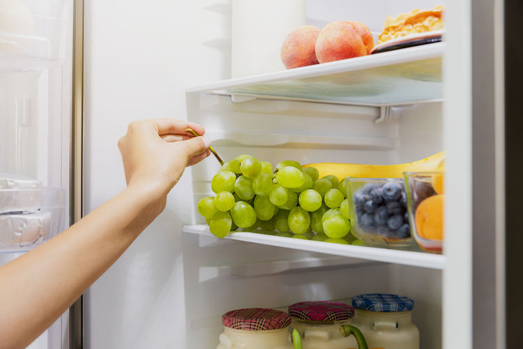 Woman hand taking, grabbing or picks up green bunch of grapes out of open refrigerator shelf, fridge