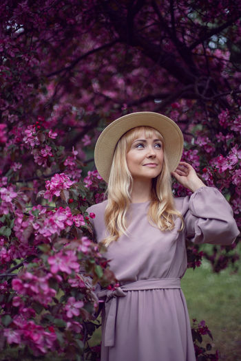 Portrait young beautiful woman in a pink dress and hat stands by a blooming pink apple tree