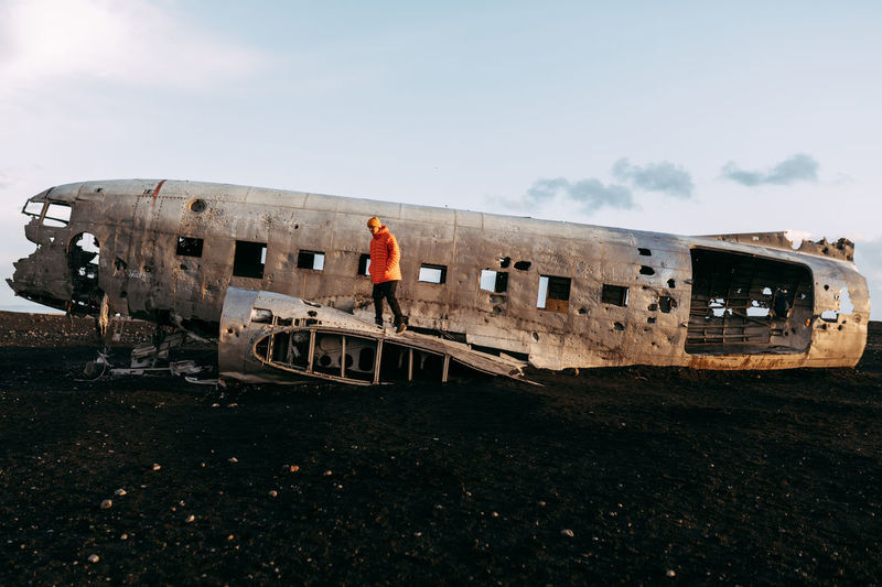 Side view of young tourist standing on wrecked aircraft between deserted lands and blue sky