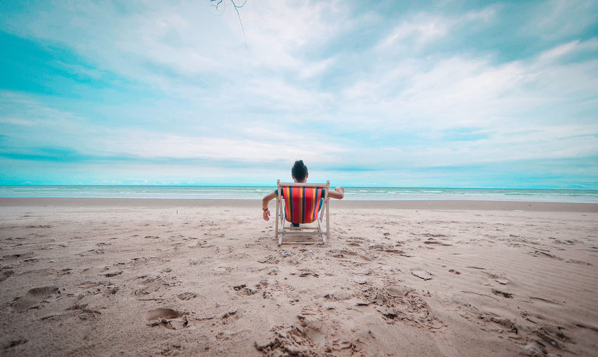 Rear view of man relaxing on beach against sky
