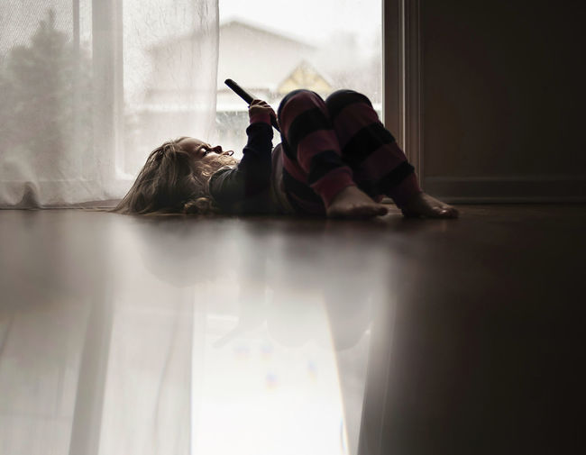 Girl using tablet computer while lying on floor against curtain at home