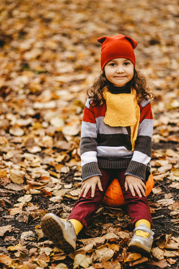 Happy cute little girl child in warm bright clothes sit on a pumpkin in the autumn forest outdoors