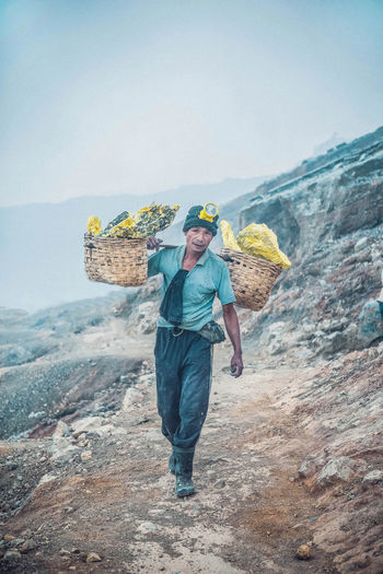 Full length of man carrying sulhpur rocks in basket against sky