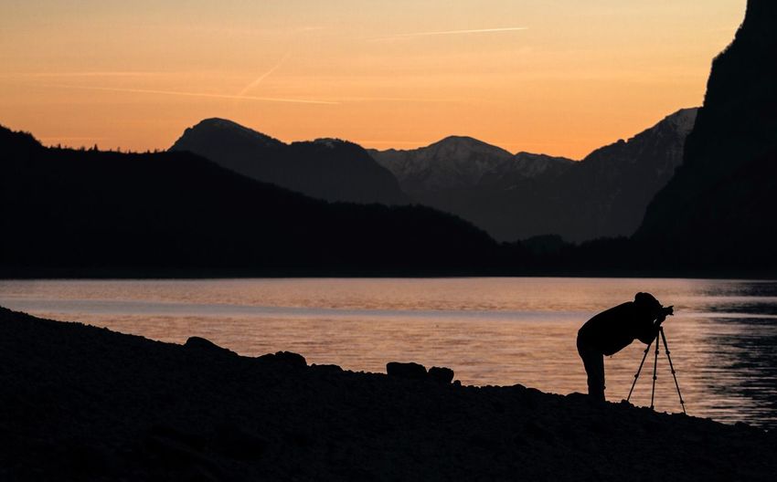 Silhouette man photographing lake against sky during sunset