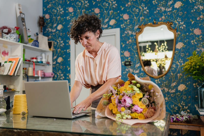 Concentrated female florist using netbook while standing at table and working in flower store
