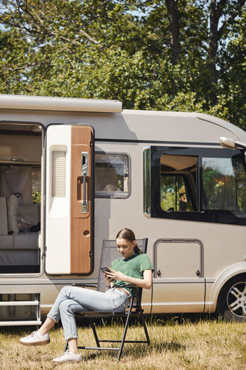 Teenage girl using mobile phone while sitting on folding chair by camper van