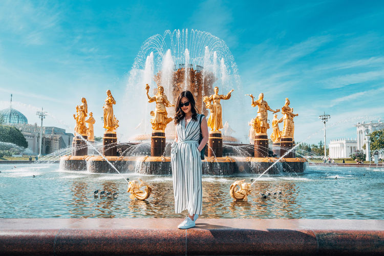 Woman standing by fountain in city against sky