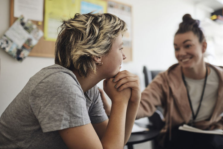 Smiling mental health professional looking at female student sitting with hand on chin in school office