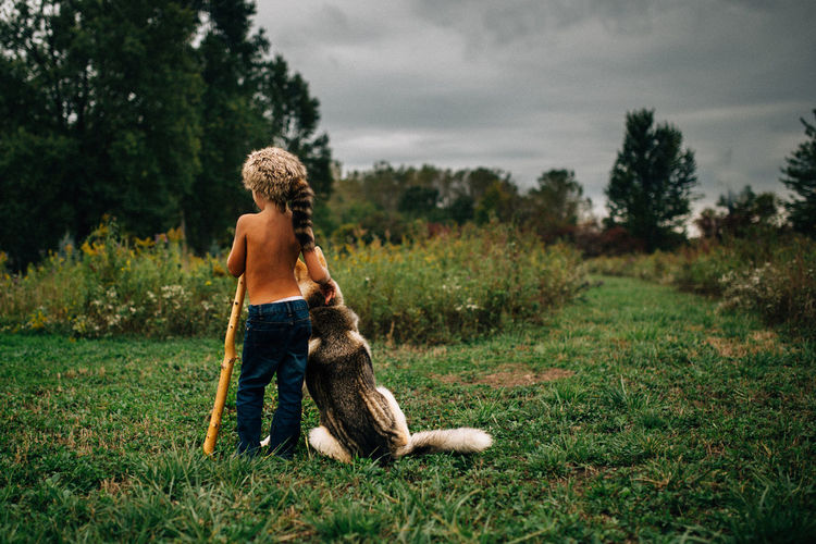 Little boy wearing coon skin hat standing with dog in a field