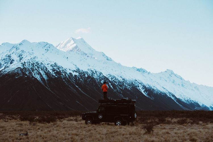 Person standing on top of car against snowcapped mountains