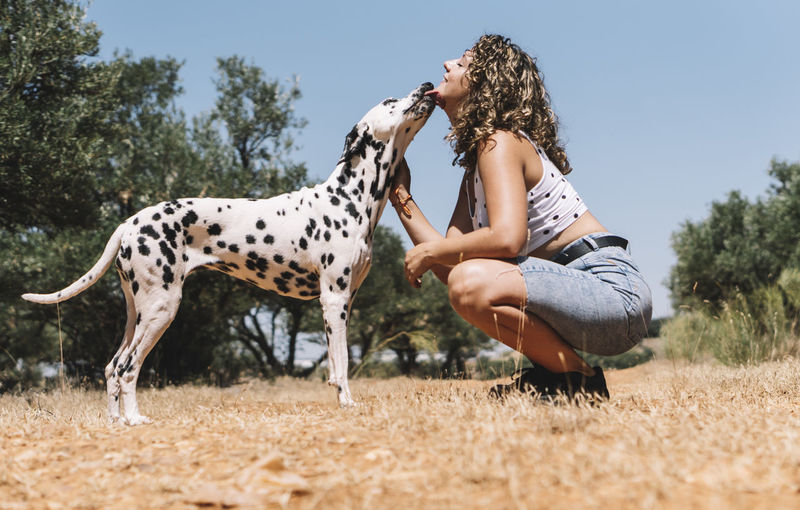 Dog licking face of young woman crouching on field during sunny day