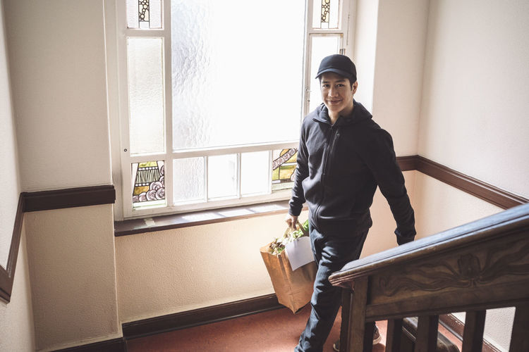 High angle portrait of smiling delivery man with package against window