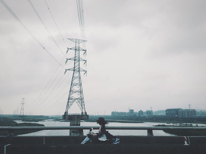 Woman riding motor scooter on bridge by electricity pylon against cloudy sky
