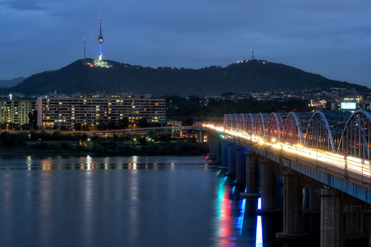Illuminated buildings by river against sky at dusk