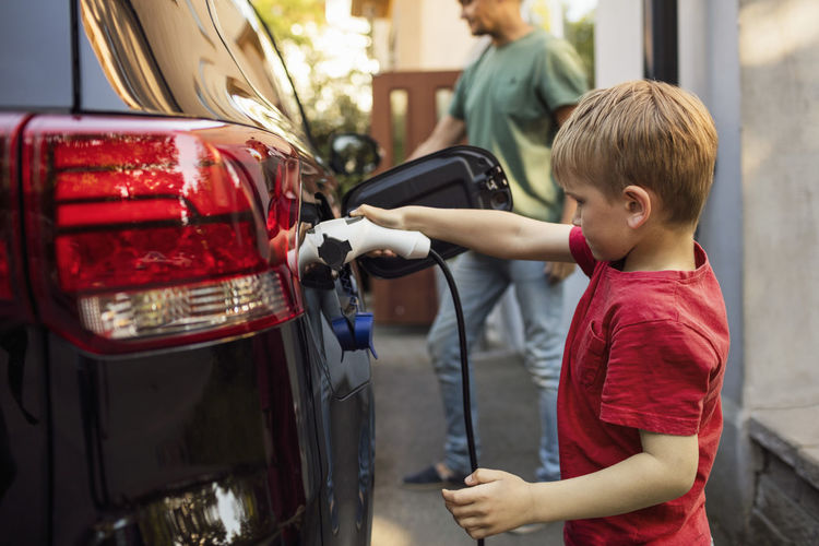 Boy charging electric car against father standing by house