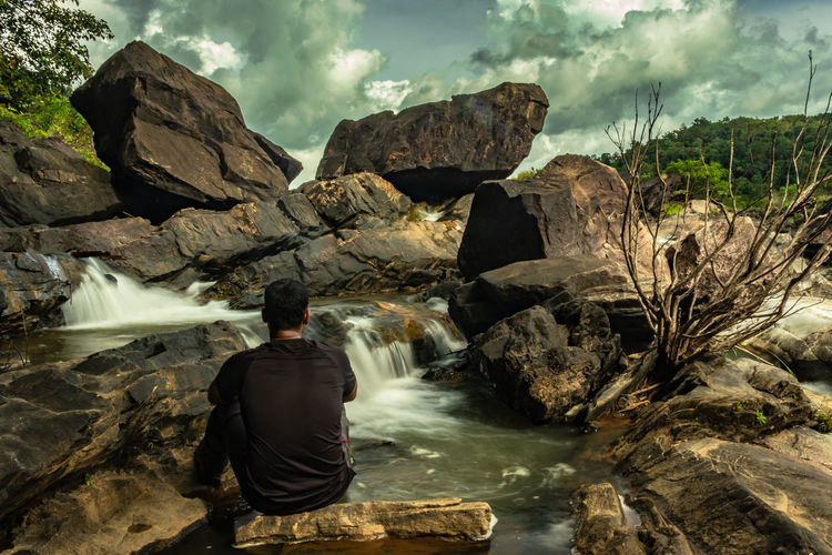 Rear view of man sitting on rock looking at waterfall