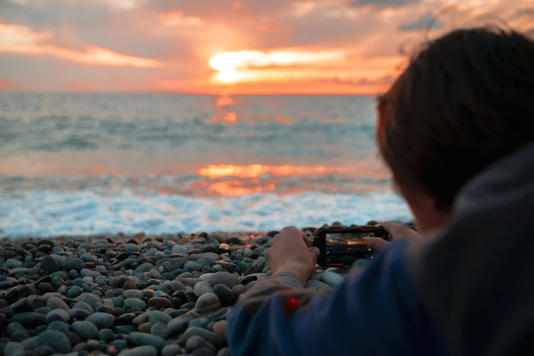 Rear view of man photographing with mobile phone at beach against cloudy sky during sunset