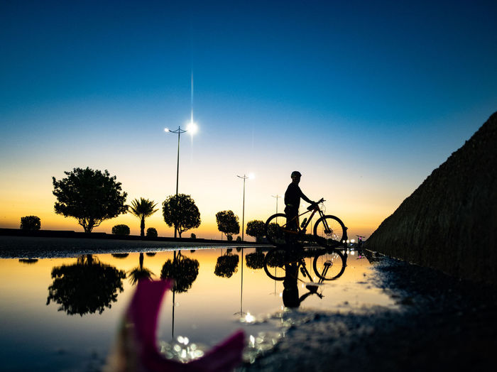 Silhouette person riding bicycle on lake against sky during sunset