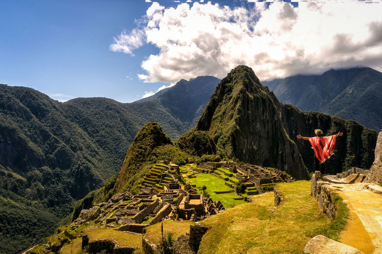 Back view of unrecognizable traveler in traditional poncho and hat admiring scenic mountainous valley while standing on viewpoint with outstretched arms during trip in machu picchu person
