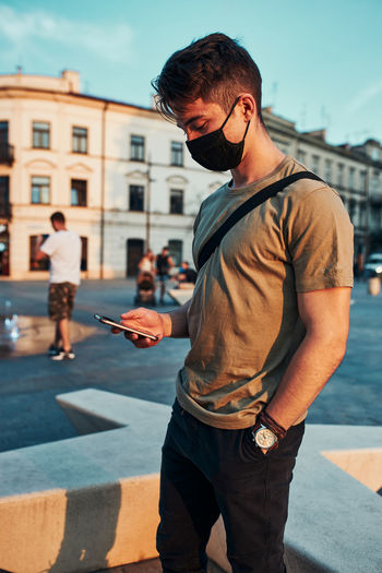 Young man standing in the city center in the evening, using smartphone, wearing the face mask