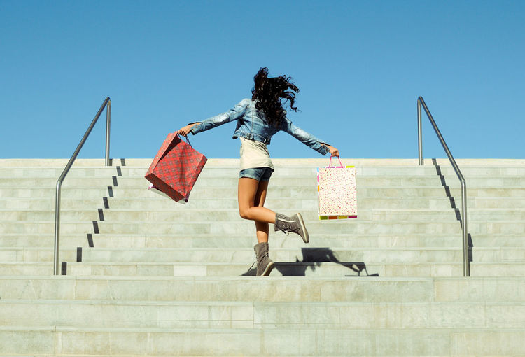 Low angle view of mid adult woman with shopping bags moving up on steps against clear blue sky