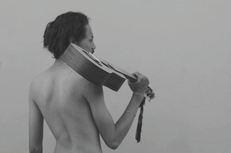 Rear view of shirtless man holding guitar while standing against clear sky