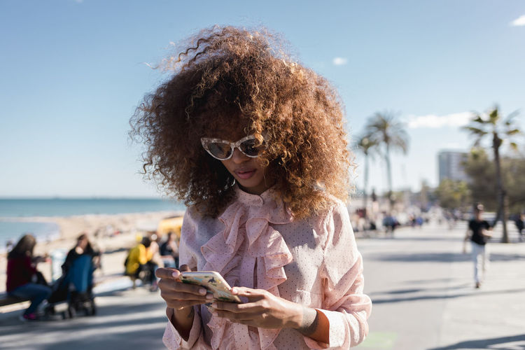 Stylish young woman using cell phone at seaside promenade