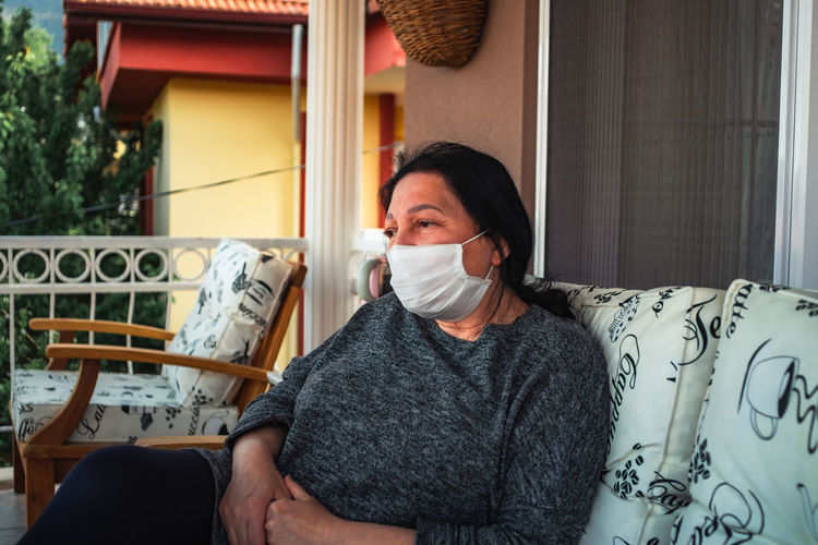 Middle aged caucasian woman with medical face mask, resting on cpuch on veranda during covid threat.