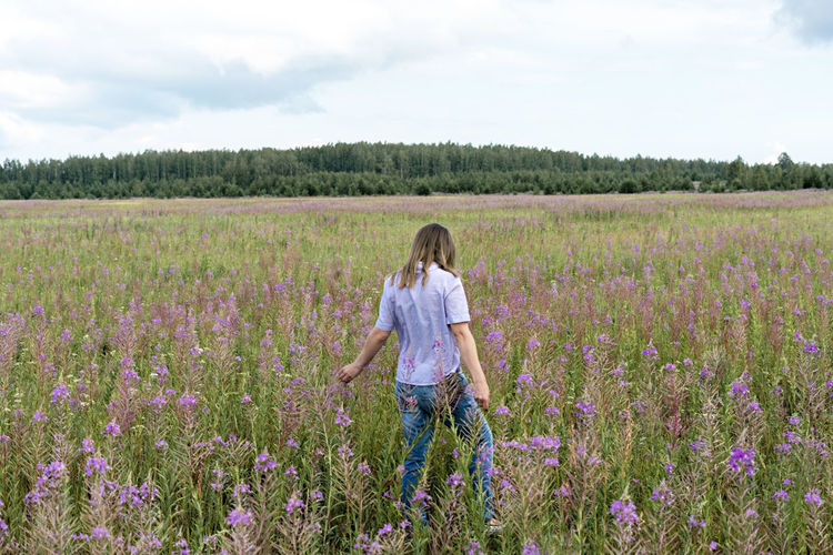 Young beautiful blond woman in purple shirt from behind walking in meadow among flowers fireweed 