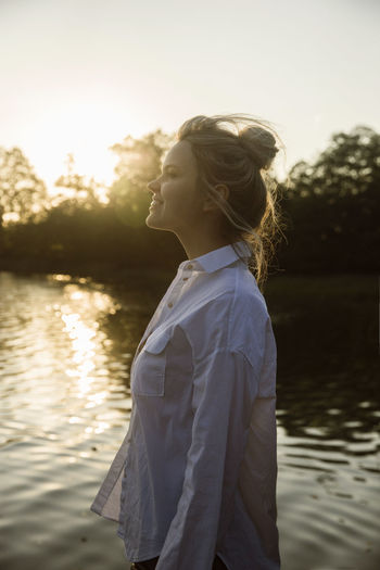 Side view of woman standing by lake against sky during sunset