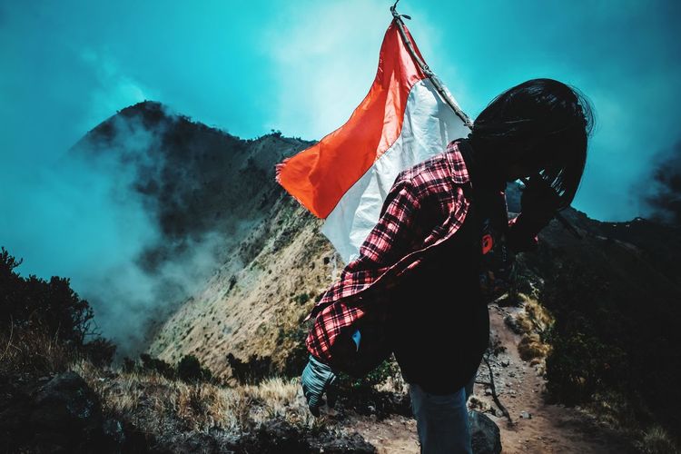 Woman holding flag while standing on mountain against sky