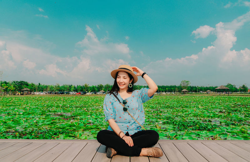Full length of woman wearing hat sitting on pier against agricultural farm