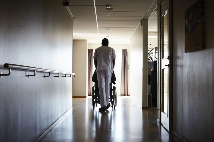 Full length rear view of male nurse pushing senior man on wheelchair at hospital corridor