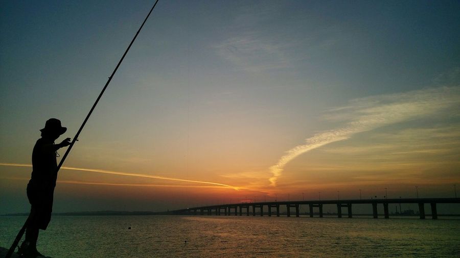 Silhouette man fishing in sea against sky during sunset