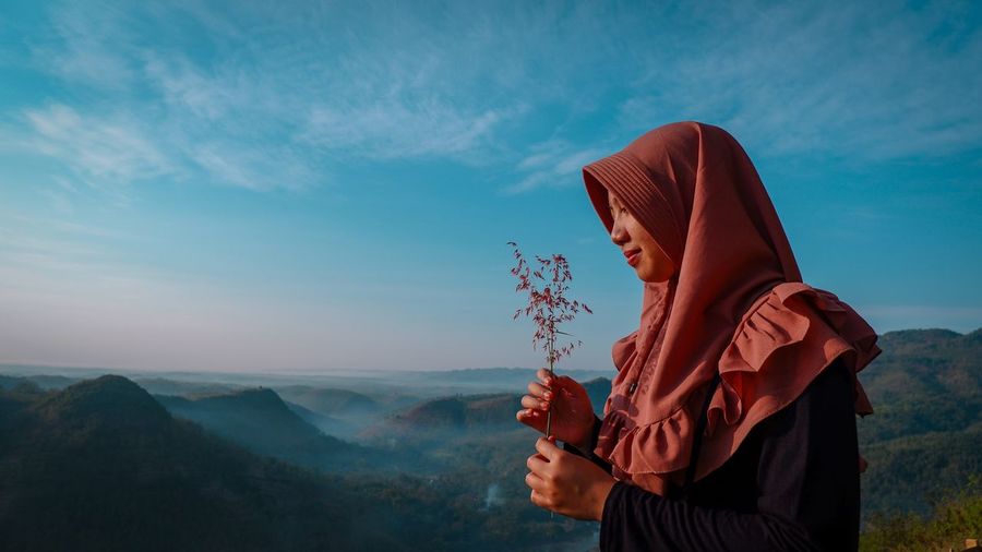 Side view of woman wearing headscarf while holding plant on mountain against sky