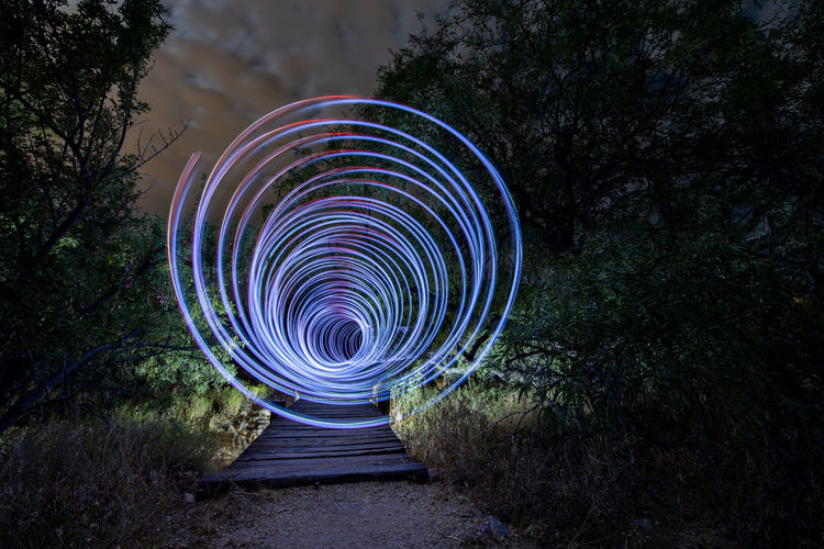 Spiral light trails on wooden path amidst desert trees at night. 