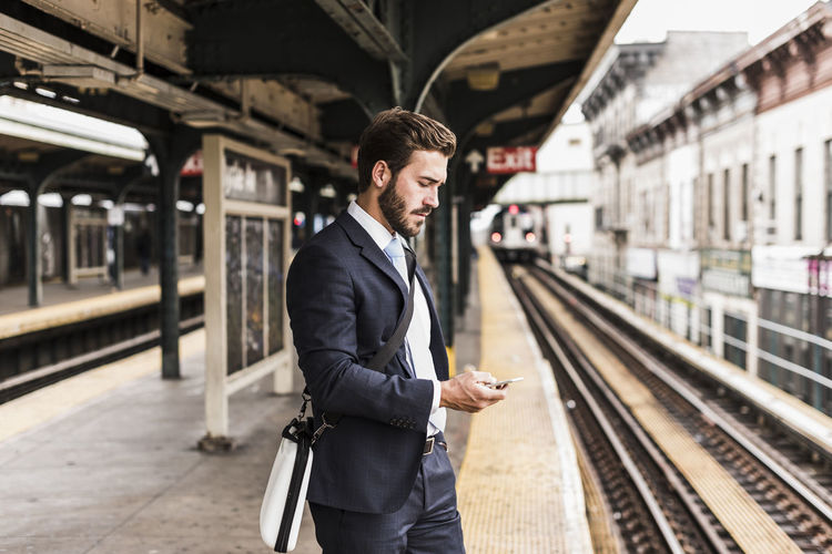Young businessman waiting at metro station platform, using smart phone